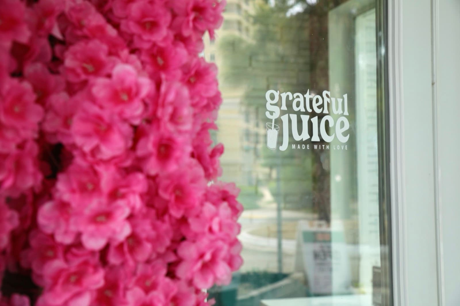 The Grateful Juice window decal seen past a wall of bright pink flowers