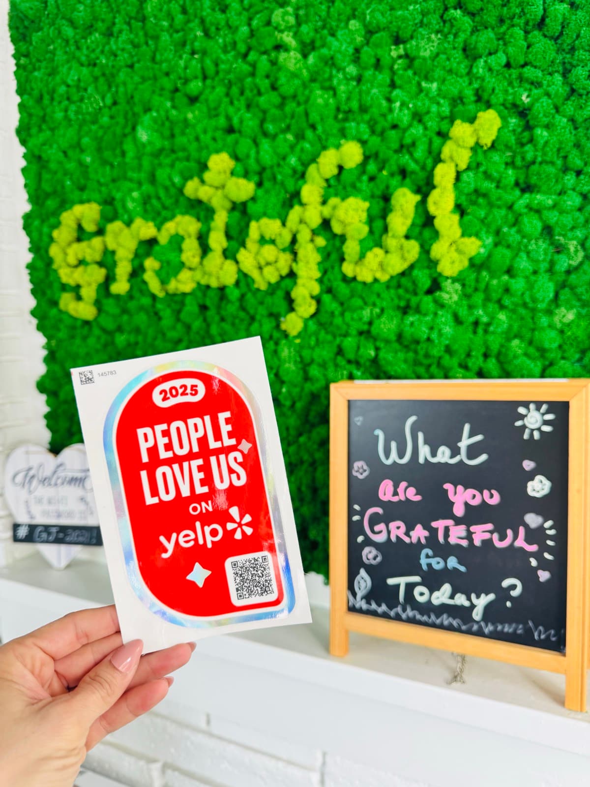 The 2025 People Love Us on Yelp sticker held up in front of the shop's mossy Grateful wall